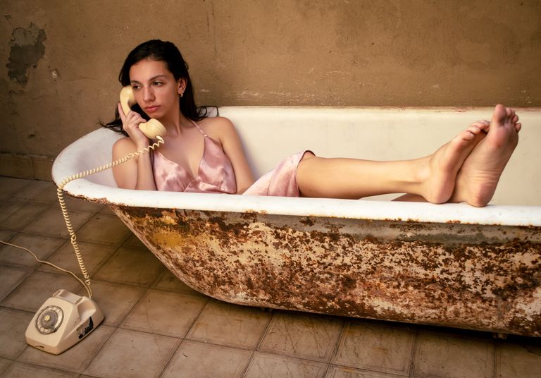 Woman in silk dress in vintage bathtub, using rotary phone. Rustic, nostalgic vibe.